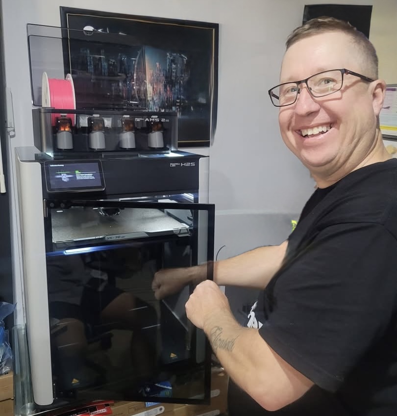 Anthony Rushby, founder of PrintIt4Me, smiling next to a 3D printer in his workshop.